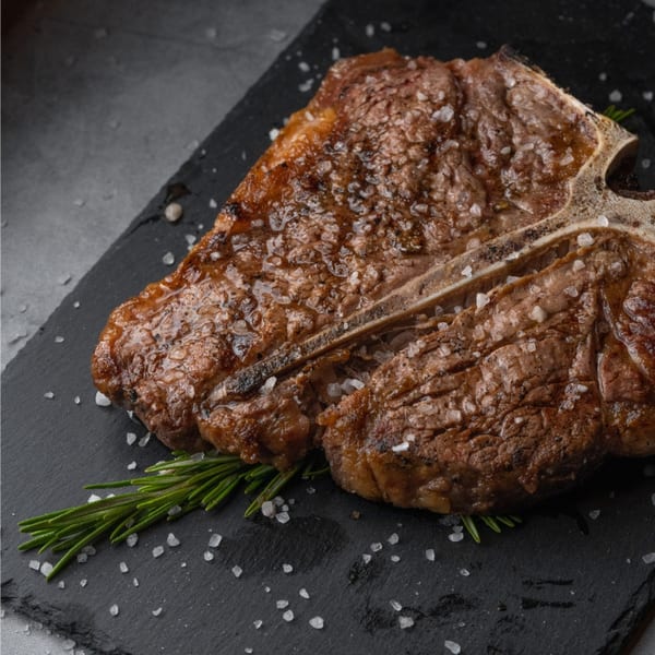 A perfectly grilled T-bone steak, seasoned with coarse salt and rosemary, on a dark slate.