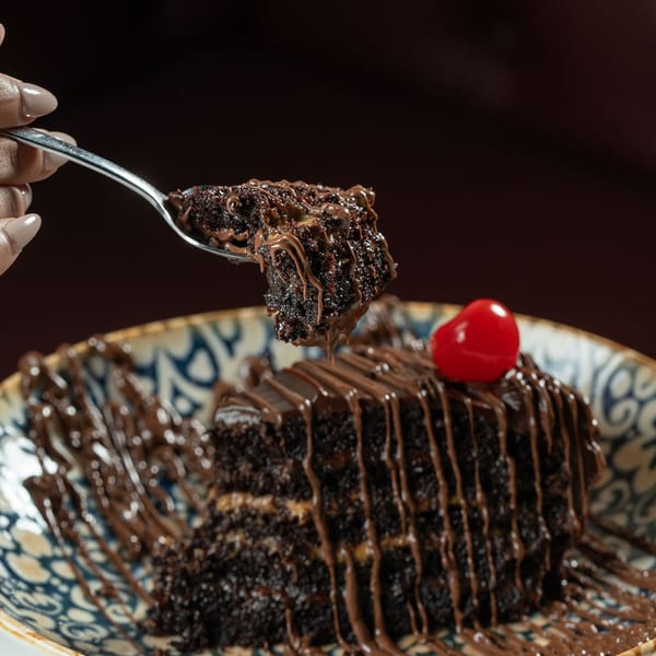 A hand holding a spoon with a bite of chocolate cake, drizzled with chocolate sauce and a cherry.