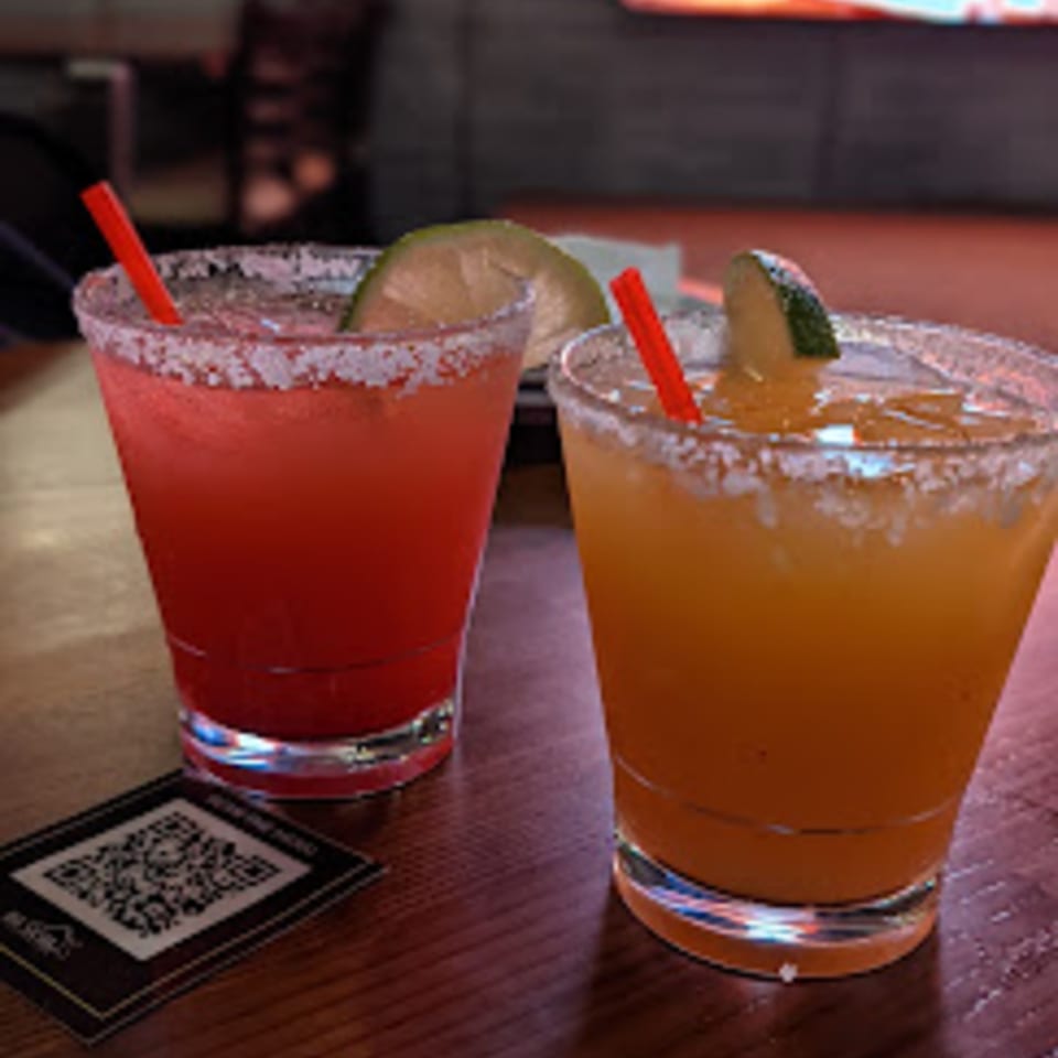 Two margaritas, one red and one orange, with salt rims, lime wedges, and red straws.