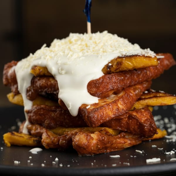 A stack of golden-brown fried plantains topped with white cream and shredded cheese on a dark plate.