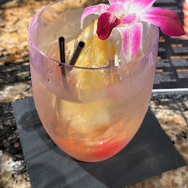 A pink cocktail with a grapefruit slice, two straws, and an orchid garnish on a dark coaster.
