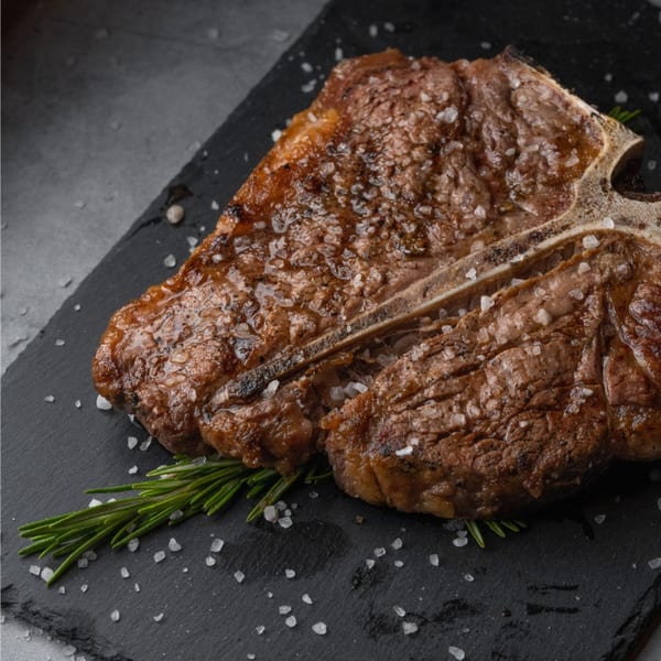 A perfectly cooked T-bone steak, seasoned with coarse salt and rosemary, on a dark slate.
