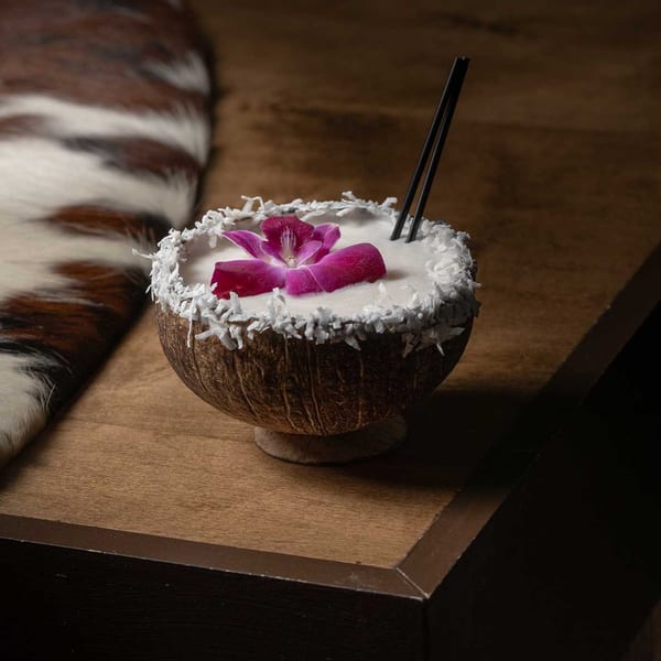 Coconut cocktail with a purple orchid and two black straws, on a wooden table with a fur rug.