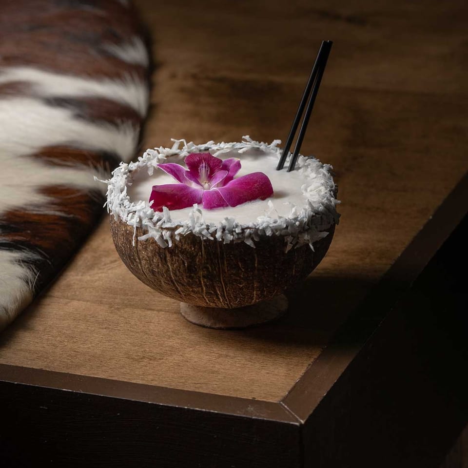Coconut cocktail with a purple orchid and two black straws, on a wooden table with a fur rug.