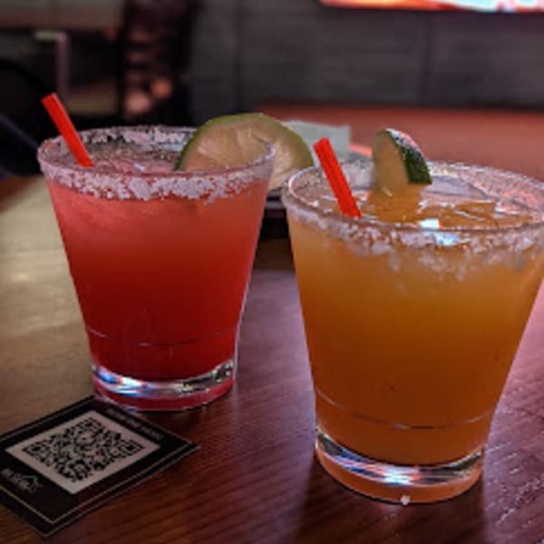Two margaritas, one red and one orange, with salt rims, lime wedges, and red straws.