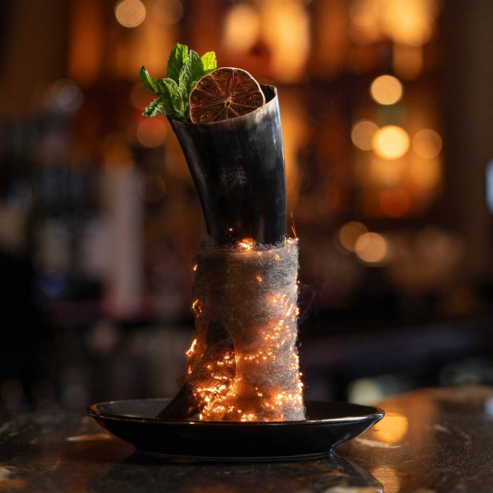 A smoking cocktail in a horn-shaped glass, garnished with mint and a dried orange slice.